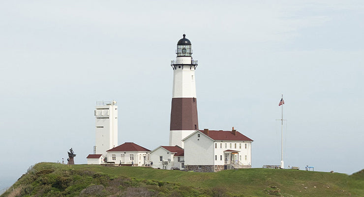 A panoramic view of the Montauk Point Lighthouse perched on a grassy cliff overlooking the ocean, under a soft overcast sky, evoking a coastal and historic charm.