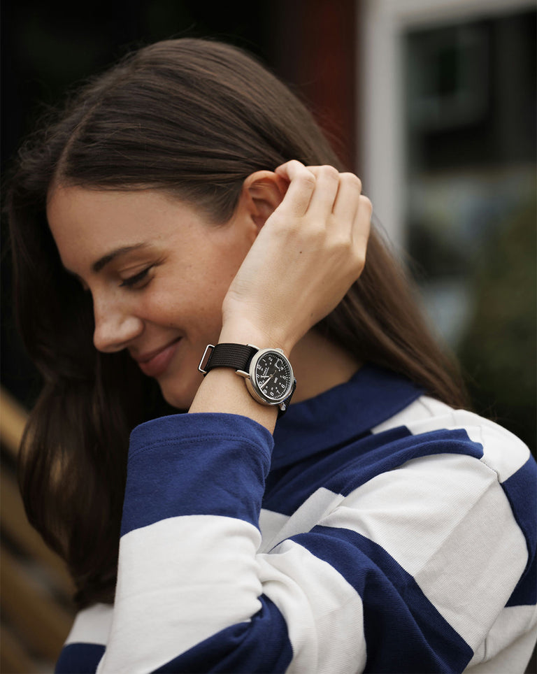 A woman smiles gently while brushing back her hair, wearing a black strap Timex field watch. She’s dressed in a navy and white striped shirt, standing in a relaxed outdoor setting.
