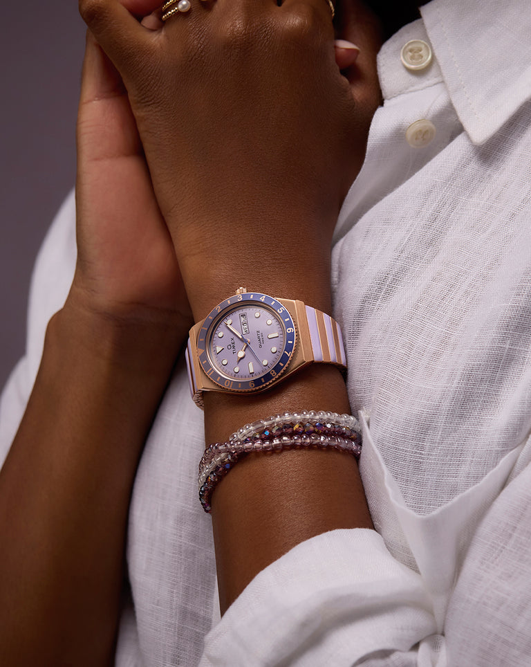 A close-up of a woman’s wrist adorned with a striking two-tone wristwatch in rose gold and lavender, featuring a blue bezel and day-date display. She is dressed in a white textured blouse, and wears layered pearl and crystal bracelets, evoking a refined and feminine look with a contemporary edge.