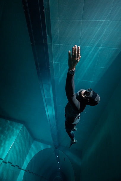 An underwater view of a freediver swimming upward in a vertical tunnel pool, fully extended with one arm reaching above. The Timex Deepwater watch is visible, highlighting its use in deep diving environments.