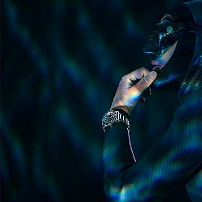 A diver in a wetsuit and mask stands partially submerged, surrounded by ambient light reflecting off the water. The Timex Deepwater watch on their wrist gleams under moody, filtered lighting, emphasizing its bold design.