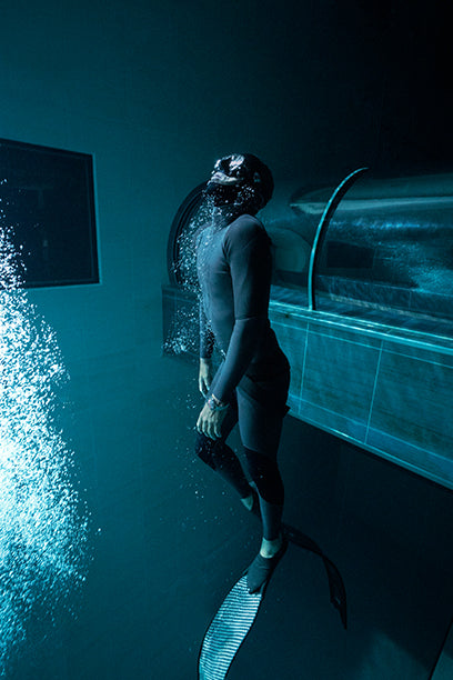 A freediver stands on a platform at the bottom of a deep dive pool, surrounded by bubbles rising to the surface. The watch on their wrist is clearly visible, reinforcing the watch’s functionality in high-pressure underwater conditions.