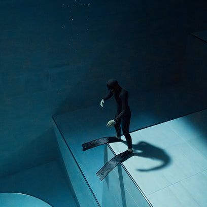 An underwater perspective of a freediver stepping cautiously onto a submerged ledge in a deep pool. The lighting casts a dramatic shadow, highlighting the diver’s form and the durability of the Timex Deepwater watch worn on their wrist.