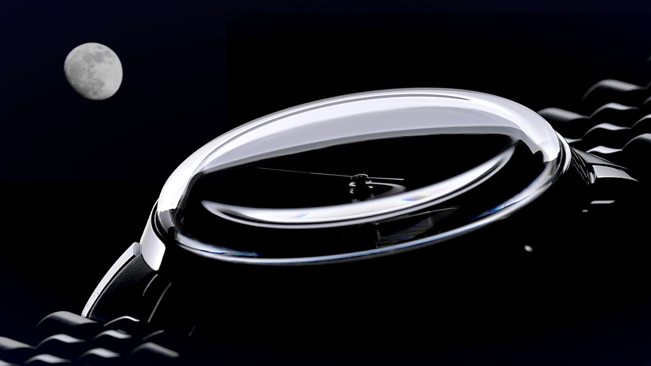 A dramatic close-up of the domed crystal on the Timex Marlin® Moon Phase watch, with a visible crescent moon in the night sky. The image highlights the curvature of the lens and the elegance of the minimalist dial beneath it.