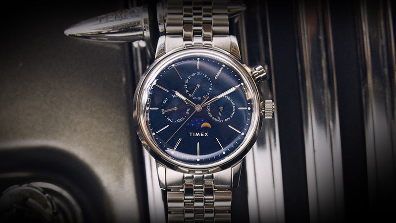 The Timex Marlin® Moon Phase is photographed upright against a classic car's chrome interior. The dial’s calendar subdials and moon phase complication are framed beautifully by the polished bracelet and elegant case.