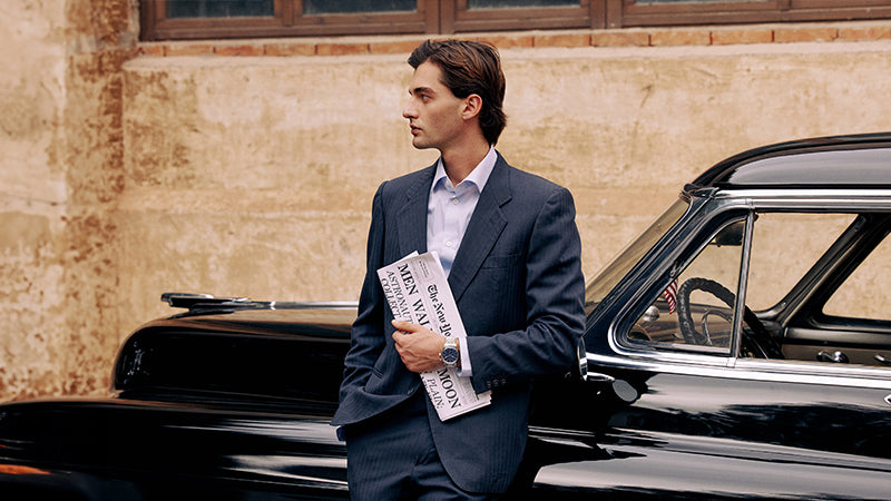 A stylish man in a dark suit leans against a vintage black car, holding a moon landing newspaper under one arm. The Timex Marlin® Moon Phase on his wrist complements the refined, retro mood of the scene.