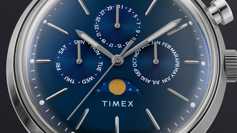 Macro view of the Timex Marlin® Moon Phase dial. The golden moon disc, calendar subdials for day, date, and month, and slender indices stand out against the deep blue background, showcasing horological precision.