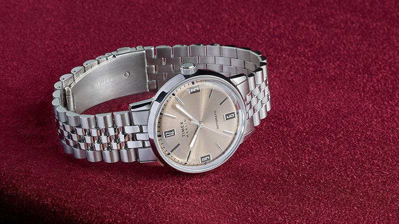 The Timex Marlin® Automatic watch is displayed on a rich burgundy fabric surface. The watch features a brushed champagne dial with minimalistic hour markers, bold black numerals at 12, 3, 6, and 9, and a polished stainless steel case and bracelet, highlighting timeless elegance and vintage design.