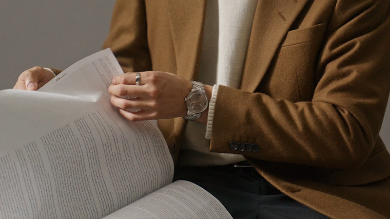 A person dressed in a camel blazer and cream sweater flips through a printed document while wearing the Timex Marlin® Automatic. The watch’s champagne dial and polished bracelet add a touch of sophistication to the casual-luxury look.
