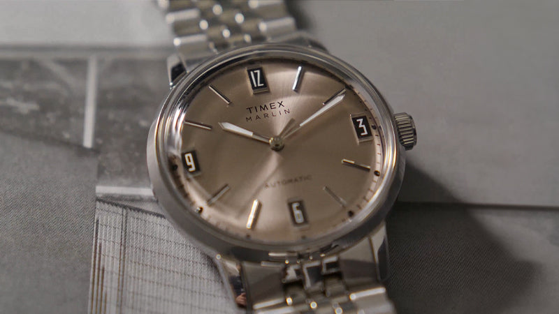 Close-up of the Timex Marlin® Automatic resting on an open magazine. The warm metallic dial and polished steel bracelet are complemented by the monochromatic tones of the background, highlighting the watch’s understated refinement.