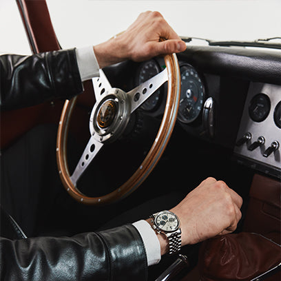 A person grips a wooden steering wheel of a vintage car while wearing the Timex Marlin® Moon Phase. The classic styling of the watch aligns perfectly with the luxurious car interior.