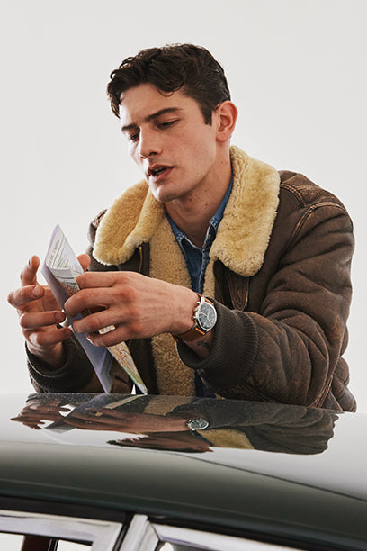 A man in a shearling jacket leans over the hood of a classic car, unfolding a road map. The Timex Marlin® Moon Phase on his wrist reinforces the spirit of exploration and heritage design.