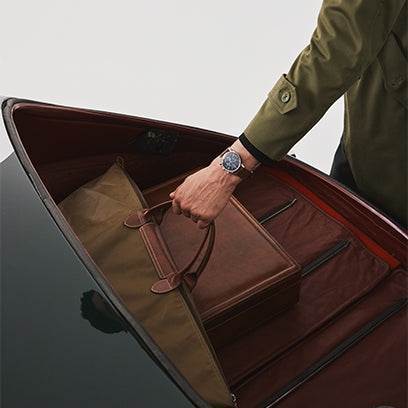 Overhead view of a man placing a leather travel bag into the open trunk of a vintage car. The Timex Marlin® Moon Phase on his wrist stands out as a symbol of style and travel-ready functionality.