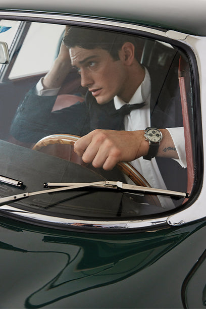 A man in formal attire, wearing a tuxedo, is seen through the windshield of a vintage green sports car, gripping the wooden steering wheel. He wears a Timex Marlin® watch on his wrist, which is prominently visible and contrasted against the crisp white shirt cuff, evoking classic elegance and automotive heritage.