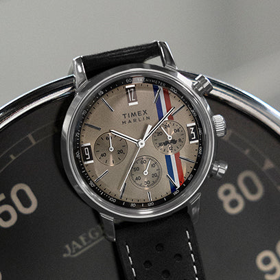 Close-up of the Timex Marlin® Chronograph placed on a vintage tachometer dial. The watch features a beige face with red, white, and blue racing stripes, three subdials, and a black perforated leather strap, capturing motorsport-inspired styling with retro flair.