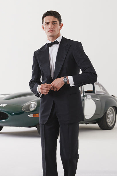 A well-dressed man stands confidently in front of a vintage racing car while adjusting his cufflinks. He wears a sleek black tuxedo and a Timex Marlin® watch on his wrist, pairing formal sophistication with racing heritage in a clean, modern setting.