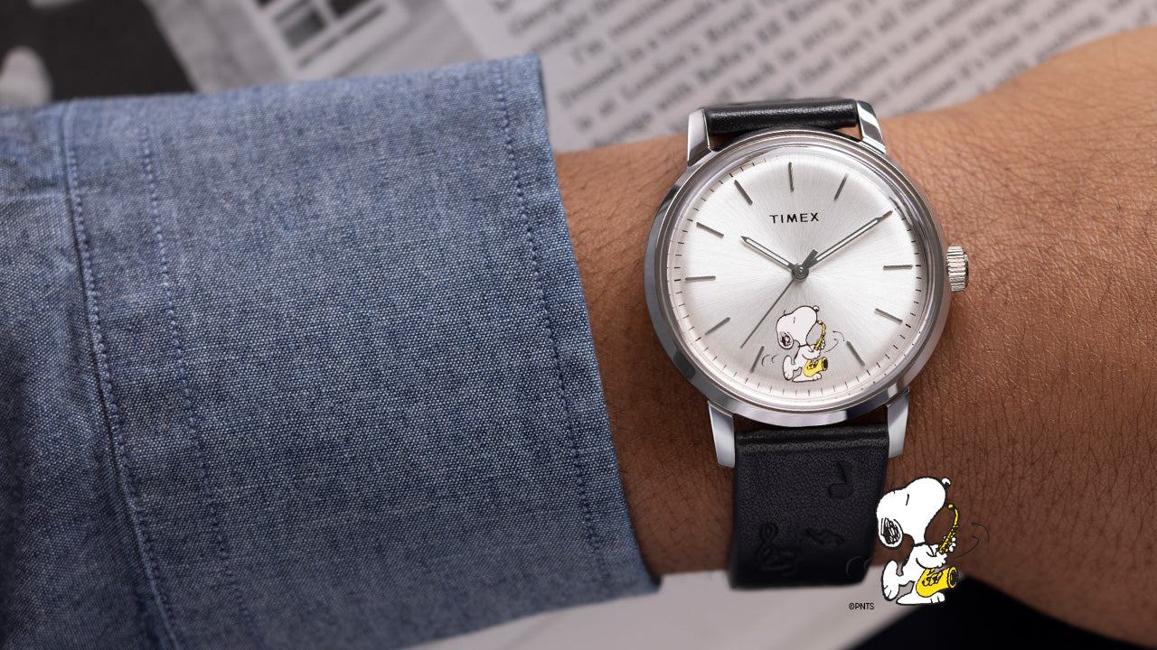 A wrist wearing the Timex Marlin® Automatic watch with a silver-tone dial featuring Snoopy playing the saxophone. The watch is paired with a black leather strap embossed with musical notes, and a chambray shirt sleeve is partially visible.
