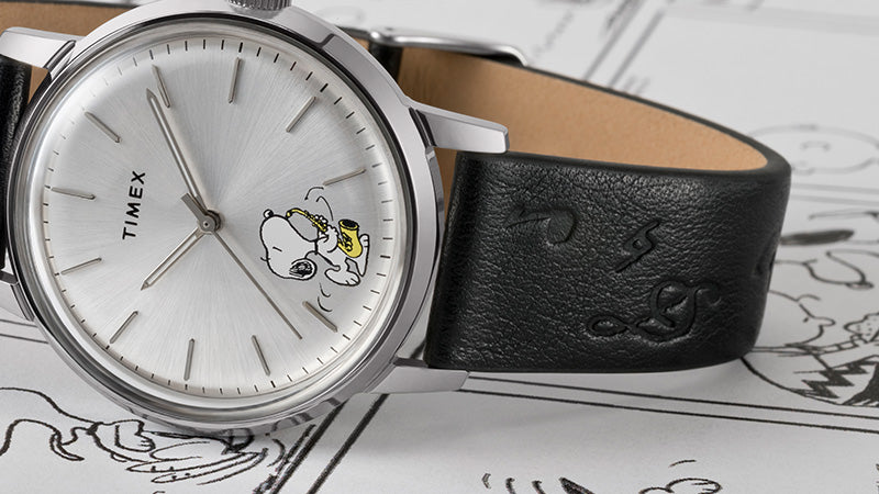 A Timex Marlin® Automatic watch with Snoopy playing the saxophone on the dial rests atop black-and-white Peanuts comic strips. The black leather strap features embossed musical notes, adding texture and theme continuity.