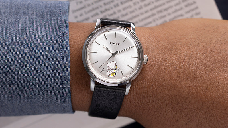 A wrist wearing the Timex Marlin® Automatic watch with a silver-tone dial featuring Snoopy playing the saxophone. The watch is paired with a black leather strap embossed with musical notes, and a chambray shirt sleeve is partially visible.