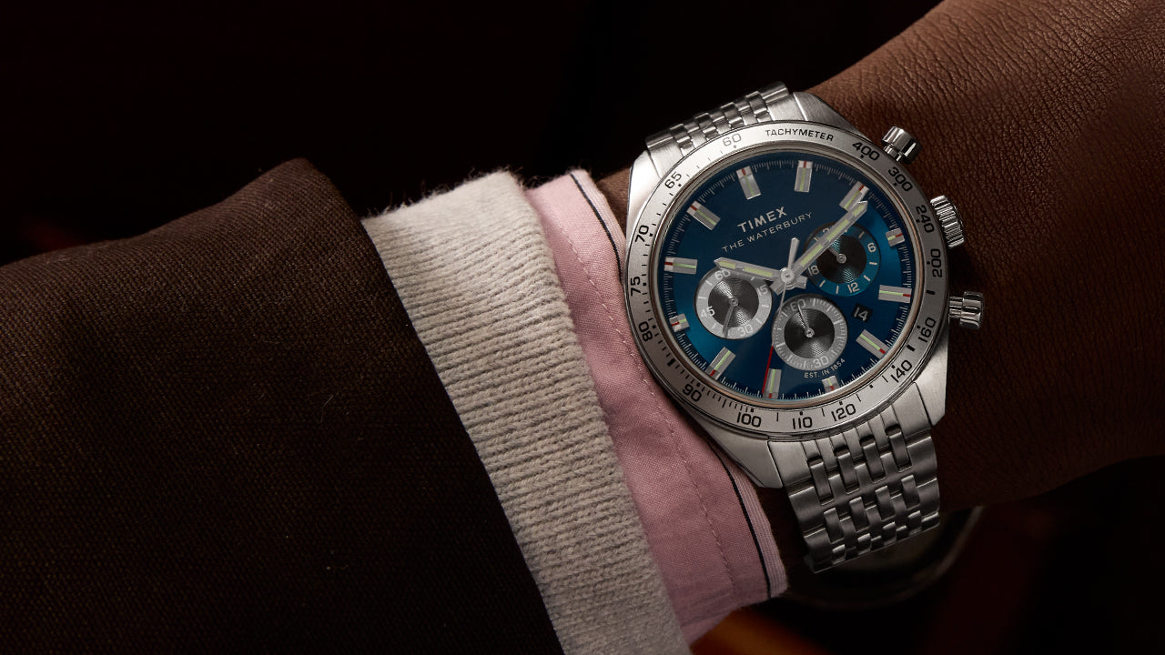 A close-up of a Timex Waterbury Heritage Chronograph watch on a person's wrist. The timepiece features a deep blue dial with three sub-dials, silver-tone hands and hour markers, and a tachymeter bezel. It is paired with a stainless steel bracelet. The wearer is dressed in a brown suit jacket, pink dress shirt, and cream corduroy sleeve, adding a touch of elegance to the scene.