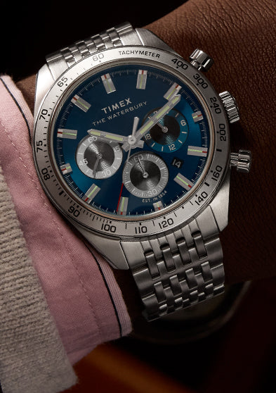 A close-up of a Timex Waterbury Heritage Chronograph watch on a person's wrist. The timepiece features a deep blue dial with three sub-dials, silver-tone hands and hour markers, and a tachymeter bezel. It is paired with a stainless steel bracelet. The wearer is dressed in a brown suit jacket, pink dress shirt, and cream corduroy sleeve, adding a touch of elegance to the scene.