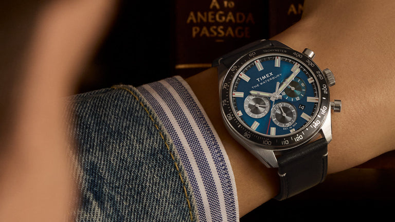 Close-up of the Timex Waterbury Chronograph on a wrist, featuring a striking blue dial, three subdials, and a tachymeter bezel. The watch is paired with a black leather strap and worn with a denim jacket and striped shirt cuff. In the background, leather-bound books—one titled “A to Anegada Passage”—add a refined, intellectual atmosphere.