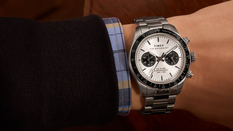 A Timex Waterbury Heritage Chronograph worn on a wrist, showcasing its stainless steel case and bracelet, black tachymeter bezel, and silver dial with black subdials. The watch is styled with a dress shirt and jacket for a refined look.