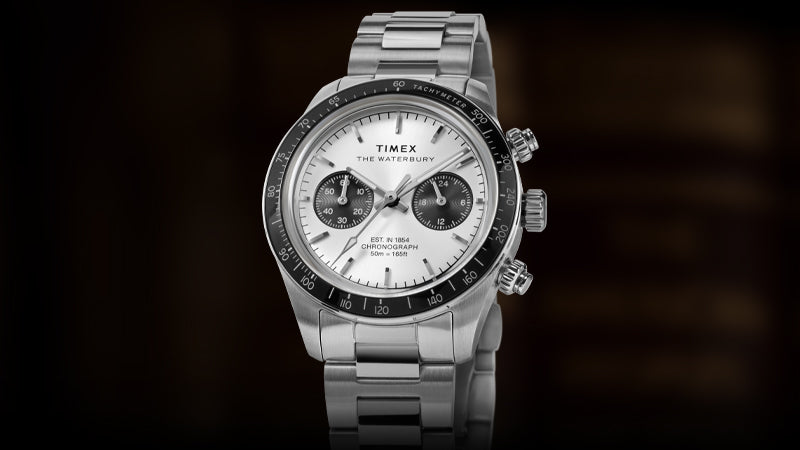 Front view of the Timex Waterbury Heritage Chronograph displaying its white dial with black chronograph subdials, tachymeter scale, and polished stainless steel bracelet against a dark background.