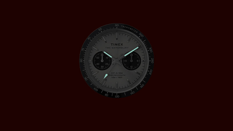 A view of the Timex Waterbury Chronograph dial in low light, showcasing its glowing luminescent hour and minute hands. The black tachymeter bezel and dual sub-dials remain subtly visible, emphasizing readability in the dark and the watch’s chronograph functionality.