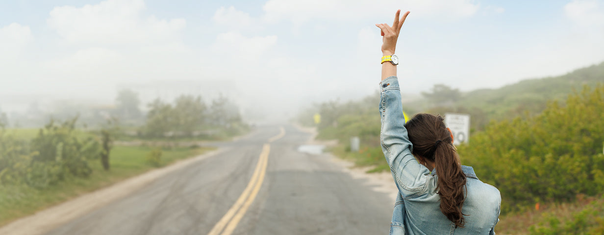 Person wearing a denim jacket and yellow watch raises their arm in a peace sign while walking down a misty country road.