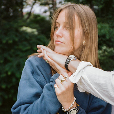 A young woman in a blue sweatshirt rests her chin on someone's outstretched arm. Both wear Timex x Dimepiece watches. She also wears pearl bracelets, gold rings, and small gold hoop earrings, gazing to the side with a relaxed expression.