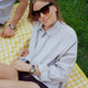 A woman wearing dark sunglasses reclines on a yellow gingham picnic blanket. She wears a lavender blouse and black shorts. The Timex x Dimepiece watch is styled with a chunky pearl bracelet and several gold rings. A man’s arm with the same watch is partly visible.