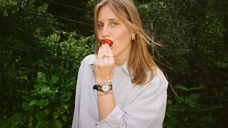 A woman stands in front of lush greenery eating a strawberry. She wears a light button-up shirt and several rings. Her Timex x Dimepiece watch is styled with layered bracelets including pearls and a braided cord.