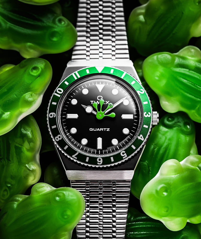 A Timex x seconde/seconde/ wristwatch with a stainless steel bracelet, featuring a black dial and a green-and-black bezel. The hour and minute hands are humorously styled as green gummy frog limbs, blending playfully with surrounding green gummy frog candies. The dial reads “QUARTZ” and prominently displays the Timex logo, enhancing the whimsical, limited-edition character of the watch.