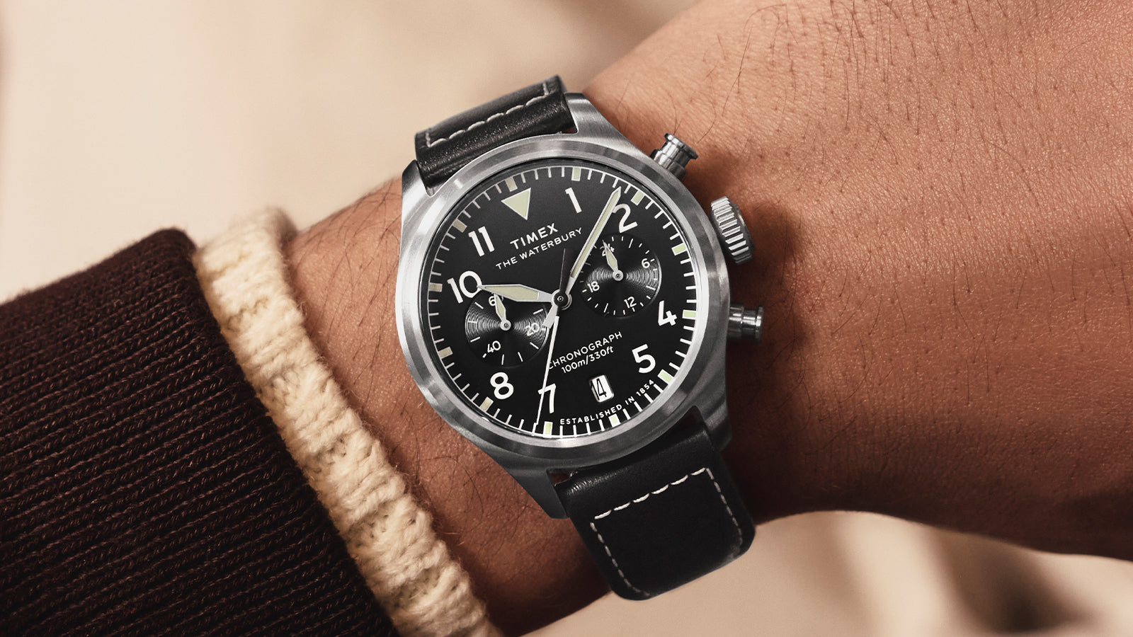 A man wearing the Timex Waterbury Ace Chronograph on his wrist, featuring a black dial with white numerals, chronograph subdials, and a black leather strap with white stitching.