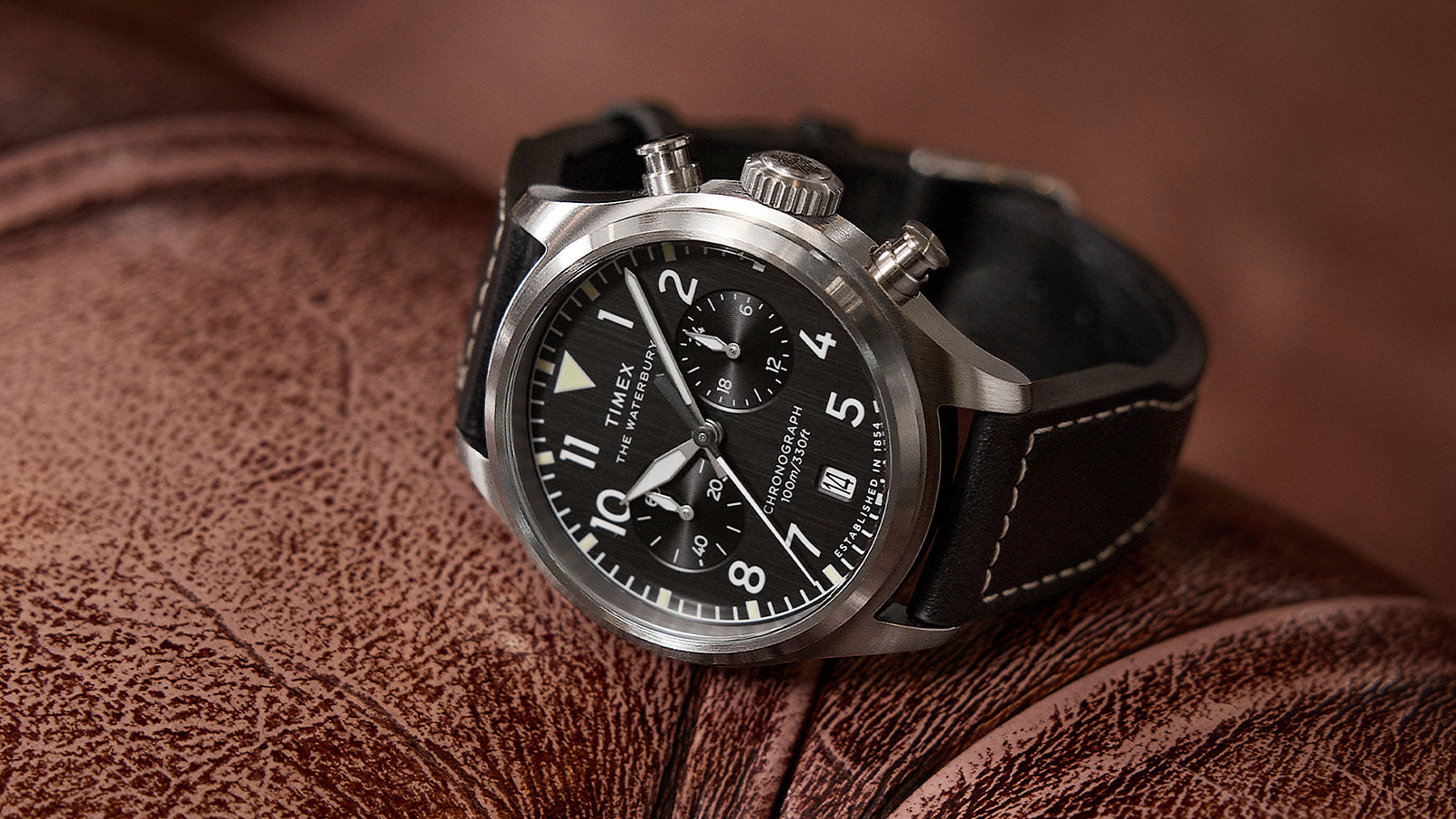 Timex Waterbury Ace Chronograph resting on a brown leather surface, showcasing its brushed stainless-steel case, black dial, chronograph subdials, and stitched leather strap.