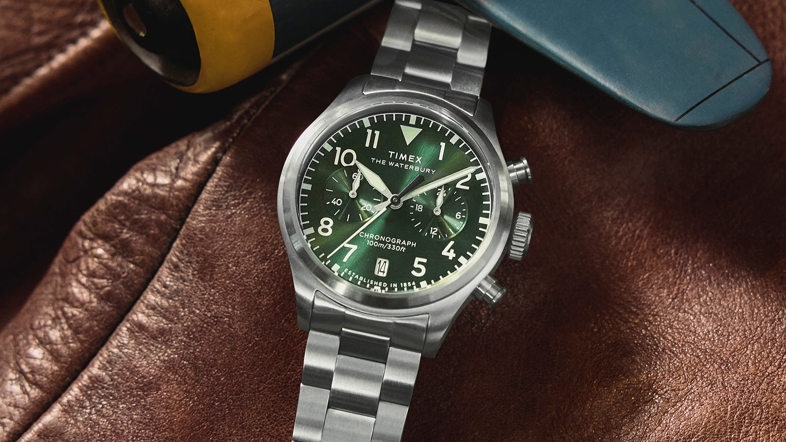 Timex Waterbury Ace Chronograph with a green dial and stainless steel bracelet resting on a brown leather surface, highlighting its brushed steel case and chronograph subdials.