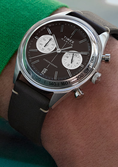 Close-up of the Timex Marlin® Jet Quartz Chronograph on wrist, featuring a rich brown dial, two contrasting white sub-dials, a date window at 6 o’clock, silver-tone case, and brown leather strap. The wearer is dressed in a green sweater.