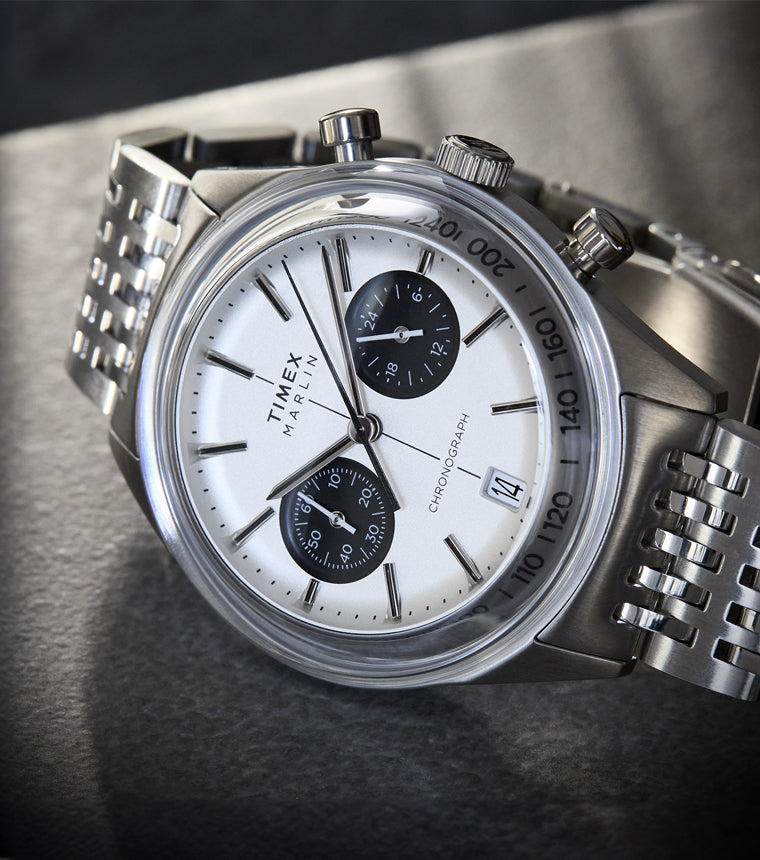 A close-up angled view of the Timex Marlin® Jet Quartz Chronograph. The watch features a white dial with two black sub-dials, a date window at 4 o’clock, silver-tone case and markers, and a polished stainless steel bracelet.