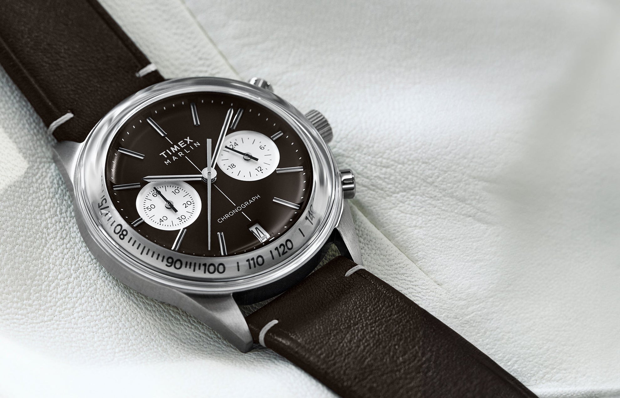 The Timex Marlin® Jet Chronograph is shown resting on a soft white backdrop. The polished case highlights the vintage-inspired brown dial and white dual sub-dials, complemented by a stitched brown leather strap.