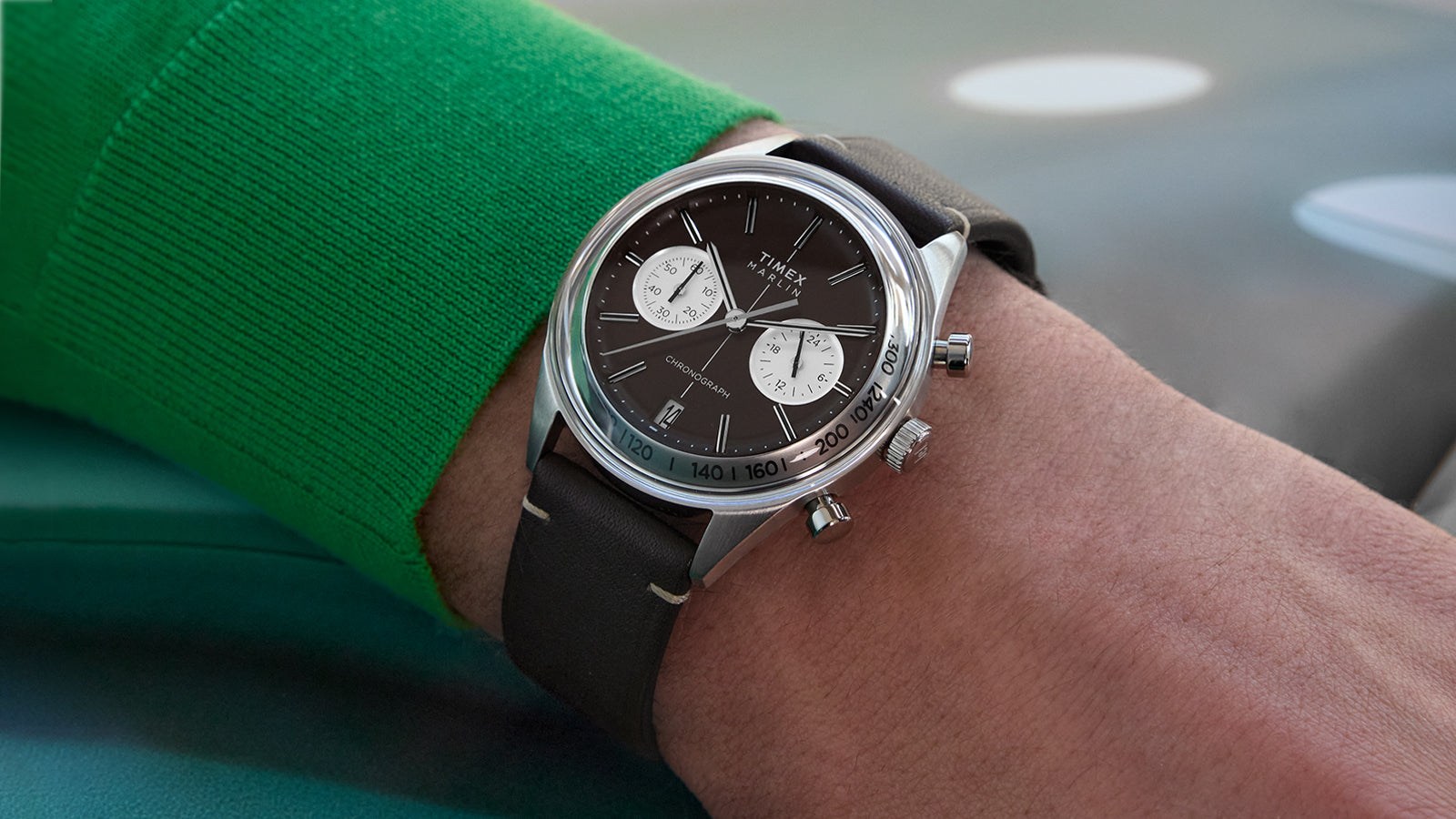 Close-up of the Timex Marlin® Jet Quartz Chronograph on wrist, featuring a rich brown dial, two contrasting white sub-dials, a date window at 6 o’clock, silver-tone case, and brown leather strap. The wearer is dressed in a green sweater.