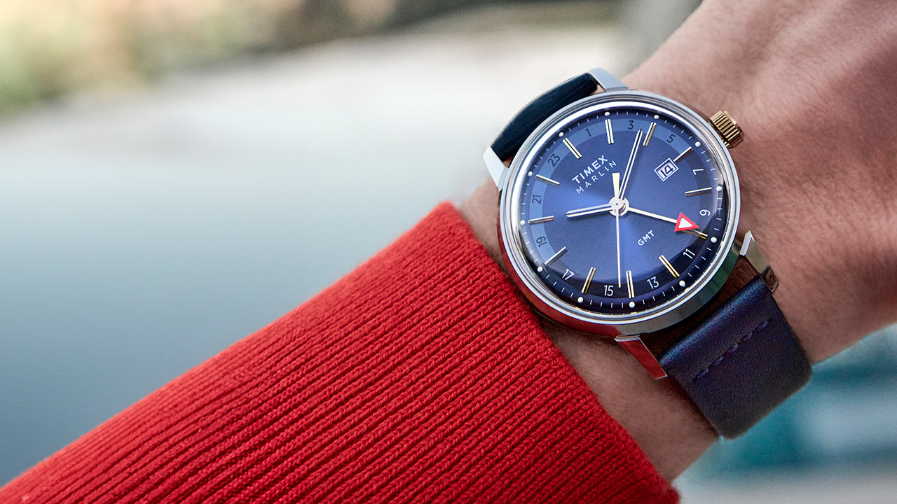 A close-up of the Timex Marlin® Quartz GMT on a wrist wearing a red sweater. The watch features a navy-blue dial, gold-tone indices and hands, a red-tipped GMT hand, and a navy leather strap with stainless steel case and gold-tone crown.