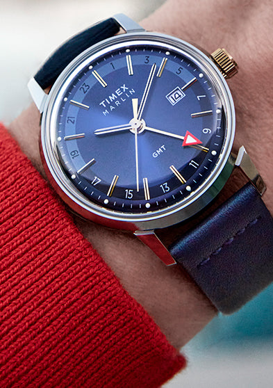 A close-up of the Timex Marlin® Quartz GMT on a wrist wearing a red sweater. The watch features a navy-blue dial, gold-tone indices and hands, a red-tipped GMT hand, and a navy leather strap with stainless steel case and gold-tone crown.