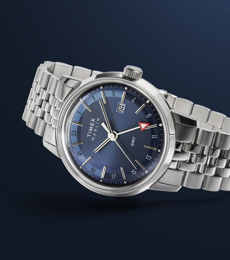 A Timex Marlin® Quartz GMT wristwatch with a deep blue sunray dial, silver-tone hour markers, a date display at 3 o’clock, and a red-arrow GMT hand. The watch has a polished stainless-steel case and a multi-link stainless-steel bracelet, shown against a dark blue gradient background.
