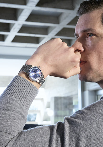 A man in a gray sweater sits in a modern setting with a pensive expression, showing off the Timex Marlin® Quartz GMT watch on his wrist. The watch features a deep blue dial with a stainless steel bracelet and red arrow GMT hand.