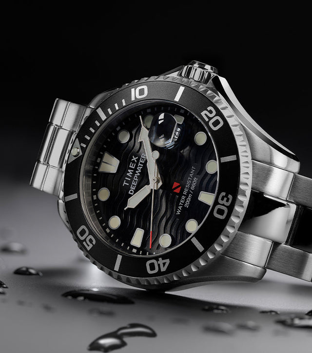 Timex Deepwater Meridian 200 dive watch in stainless steel with black rotating bezel and wavy black dial, lying at an angle with water droplets on the surface.