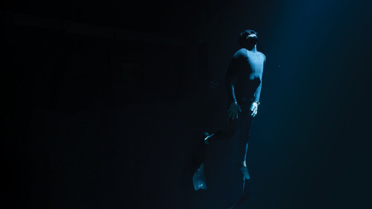 Freediver in a wetsuit illuminated by a beam of light underwater, wearing the Timex Deepwater Meridian 200 dive watch while floating upward in the deep.