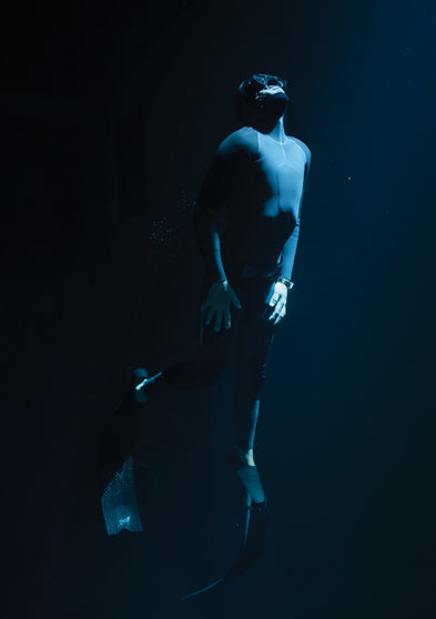 Freediver in a wetsuit illuminated by a beam of light underwater, wearing the Timex Deepwater Meridian 200 dive watch while floating upward in the deep.