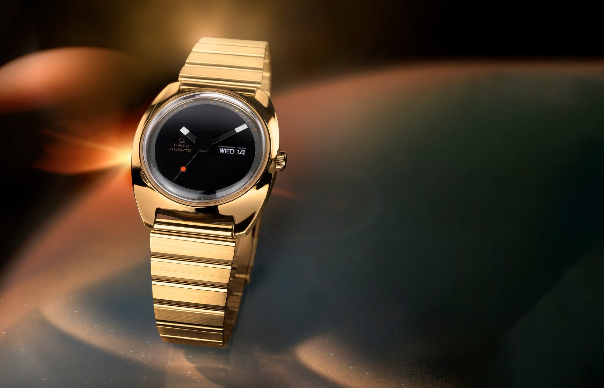 Q Timex® 1975 Enigma watch in polished gold-tone stainless steel, featuring a minimalist black dial with geometric hands, small red dot at 6 o’clock, day-date window at 3 o’clock, and integrated gold-tone bracelet, set against a warm, glowing abstract background.