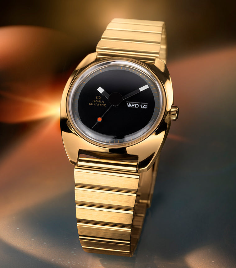 Q Timex® 1975 Enigma watch in polished gold-tone stainless steel, featuring a minimalist black dial with geometric hands, small red dot at 6 o’clock, day-date window at 3 o’clock, and integrated gold-tone bracelet, set against a warm, glowing abstract background.